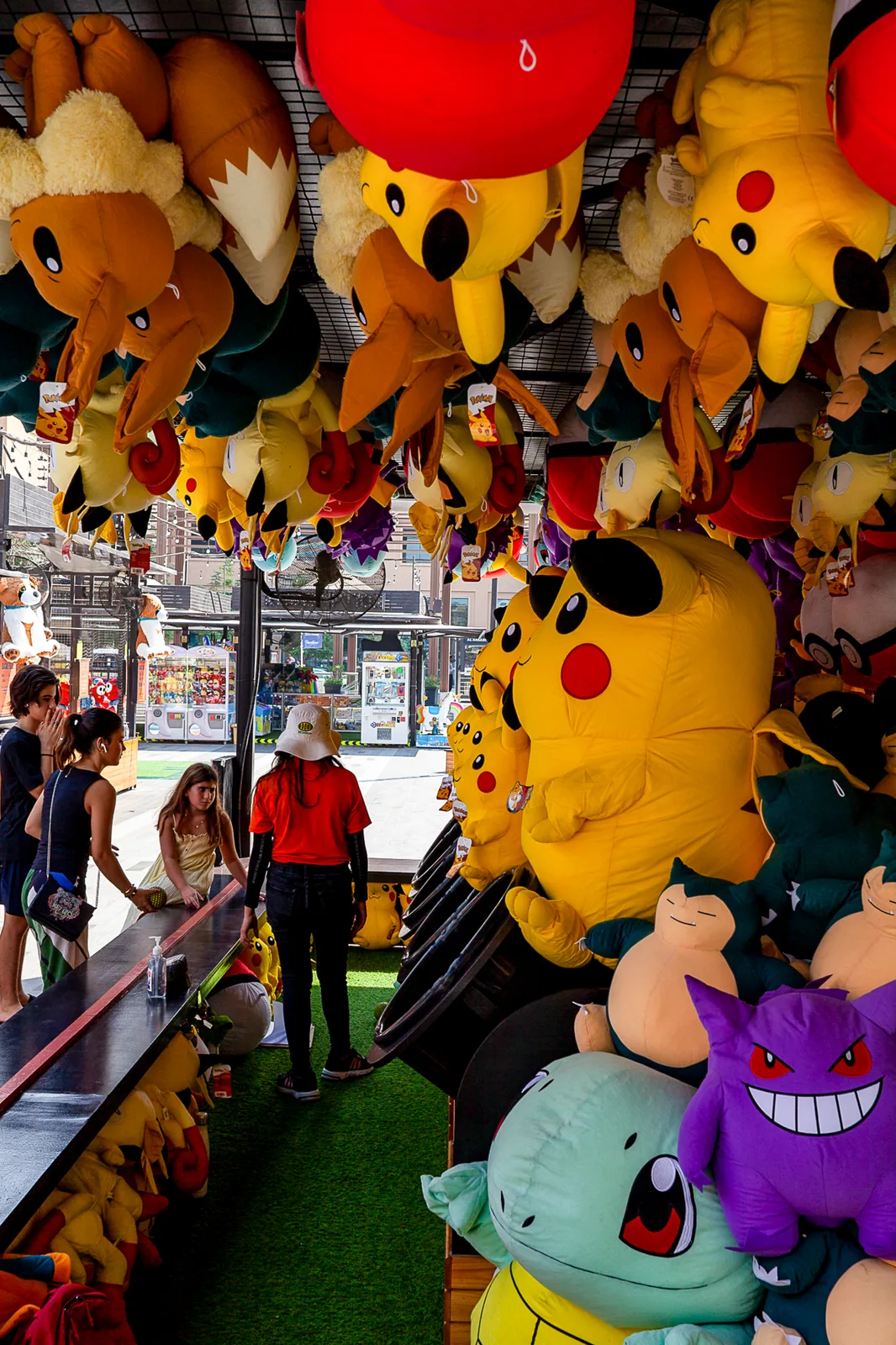Un stand de fête forain dont le plafond est couvert de grandes peluches Pokémon suspendues ; une personne en chapeau et haut rouge se tient de dos, des jeunes près du comptoir.