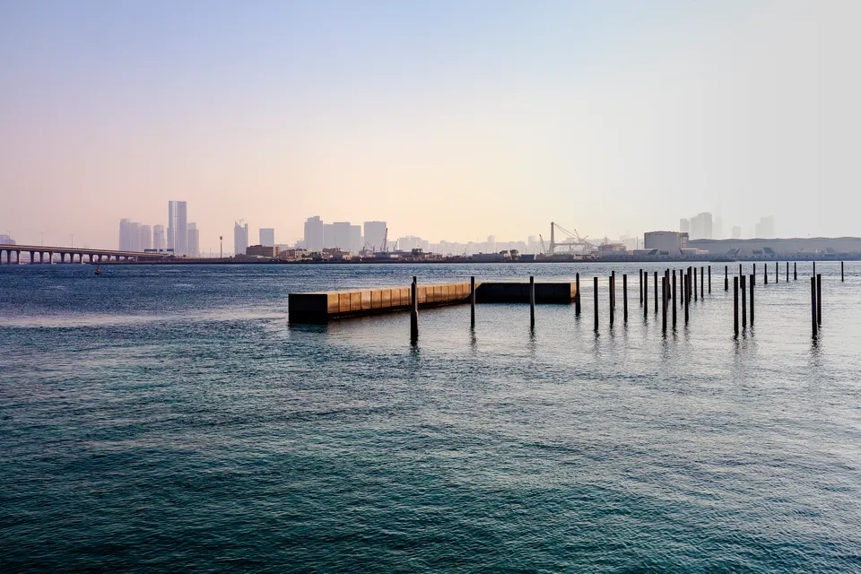 Un ponton en béton et plusieurs rangées de poteaux sombres émergent d'une mer calme, avec une skyline lointaine, un pont et des grues dans une brume pastel.