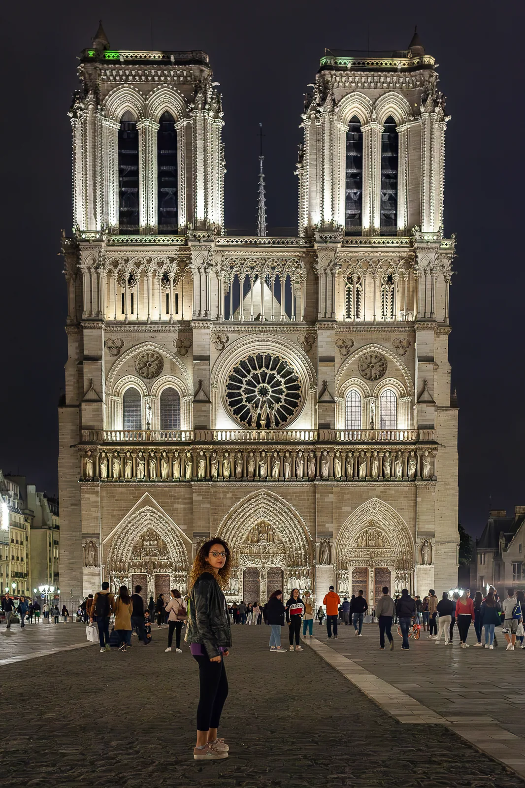 Façade illuminée d'une cathédrale gothique à deux tours, de nuit, avec rosace et portails ; au premier plan, une femme aux cheveux bouclés regarde par-dessus son épaule, des passants au fond.