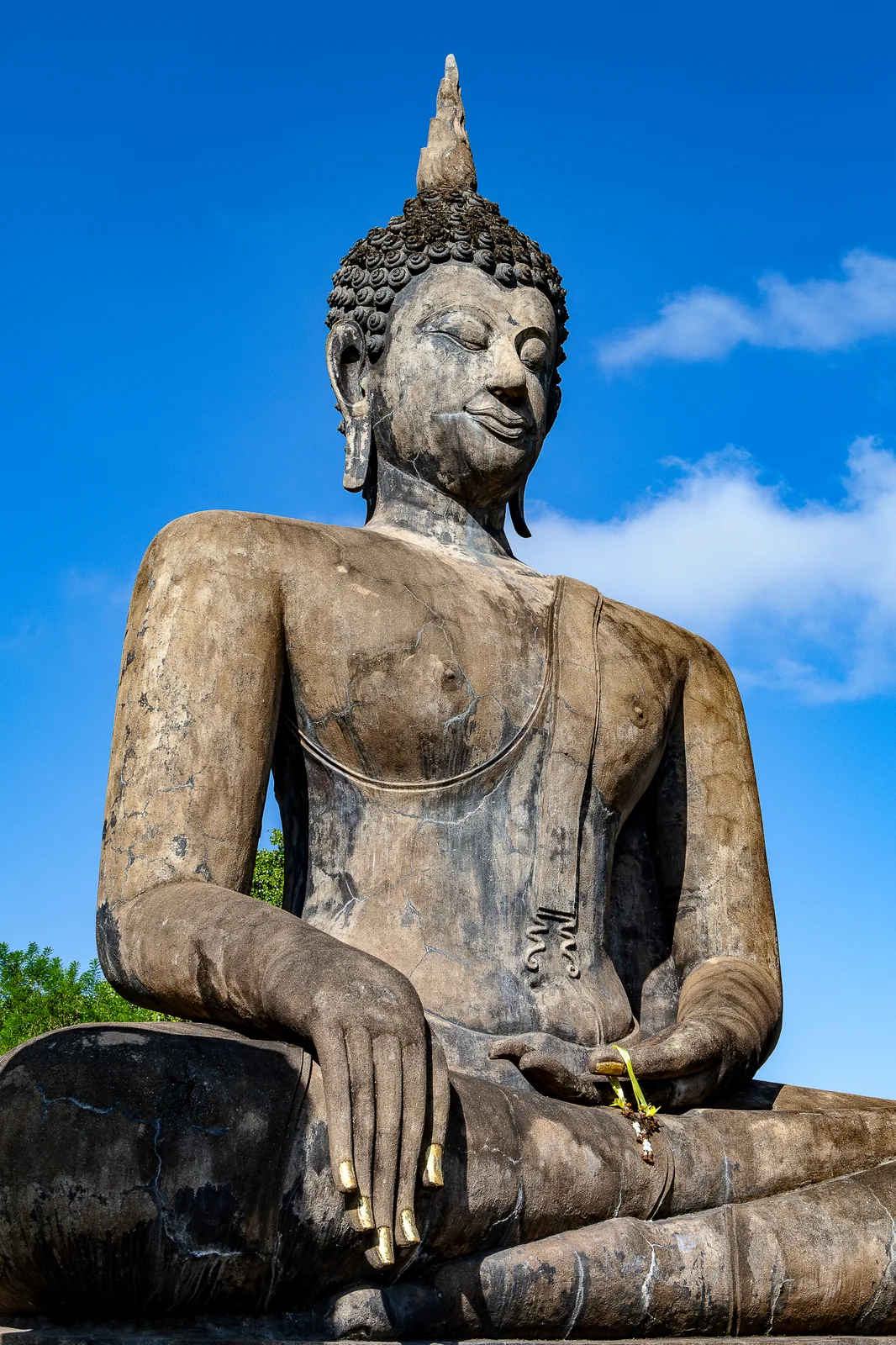 Vue en contre-plongée d'une grande statue de Bouddha assis en pierre patinée, yeux baissés, ongles dorés, une petite offrande dans la main, devant un ciel bleu avec quelques nuages.