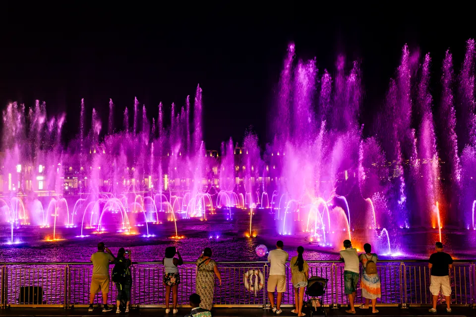 De nuit, des spectateurs vus de dos s'appuient à une rambarde face à de grandes fontaines éclairées en violet et orange, avec des bâtiments illuminés au fond.