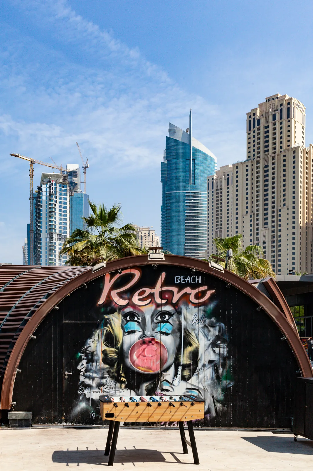 Un baby-foot en bois est posé sur une terrasse devant une fresque murale sur une façade arrondie, avec l'inscription « Retro Beach » ; des gratte-ciel et des grues se dressent derrière les palmiers sous un ciel bleu.