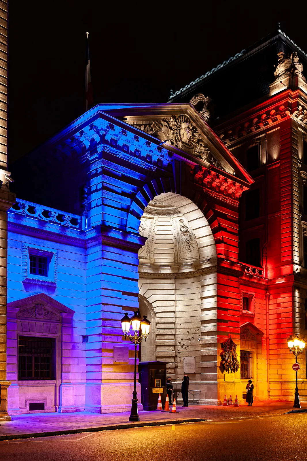 Porche monumental en pierre éclairé la nuit en bleu à gauche, blanc au centre et rouge à droite, avec lampadaires allumés, plots orange et quelques personnes devant l'entrée.
