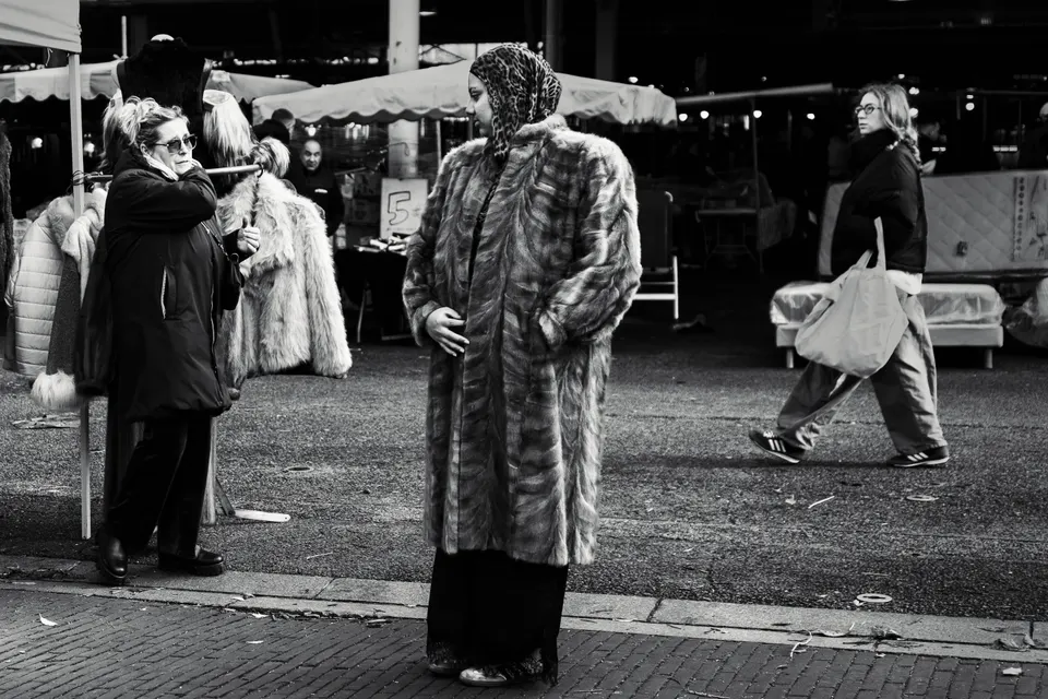 Photographie en noir et blanc d'un marché en plein air : une femme en long manteau de fourrure et foulard à motif léopard se tient debout, tandis qu'une vendeuse et une passante les entourent.