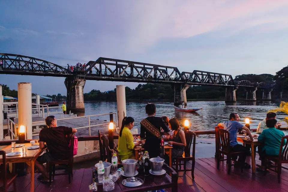 Terrasse en bois au bord de l'eau où des gens dînent à des tables éclairées par de petites lampes, face à un pont métallique et une barque sous un ciel violet.