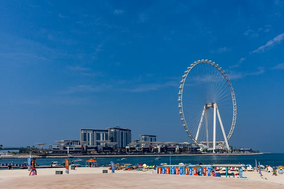 Une immense roue blanche s'élève derrière des immeubles, de l'autre côté de l'eau, face à une plage avec pédalos, parasols et quelques personnes, sous un ciel bleu.