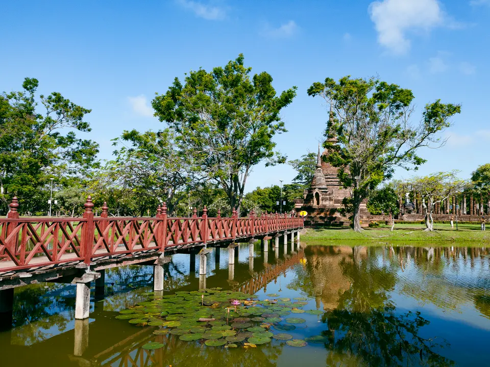 Une passerelle en bois rouge à balustrade croisillonnée traverse un étang couvert de nénuphars, vers un stupa en brique entouré d'arbres, sous un ciel bleu avec quelques nuages.