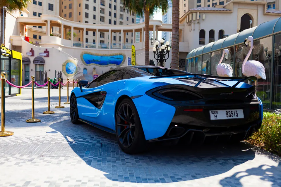 Vue arrière trois quarts d'une voiture de sport bleue et noire à plaque de Dubaï, garée sur des pavés près de potelets dorés, de deux flamants roses sculptés et d'immeubles beiges.