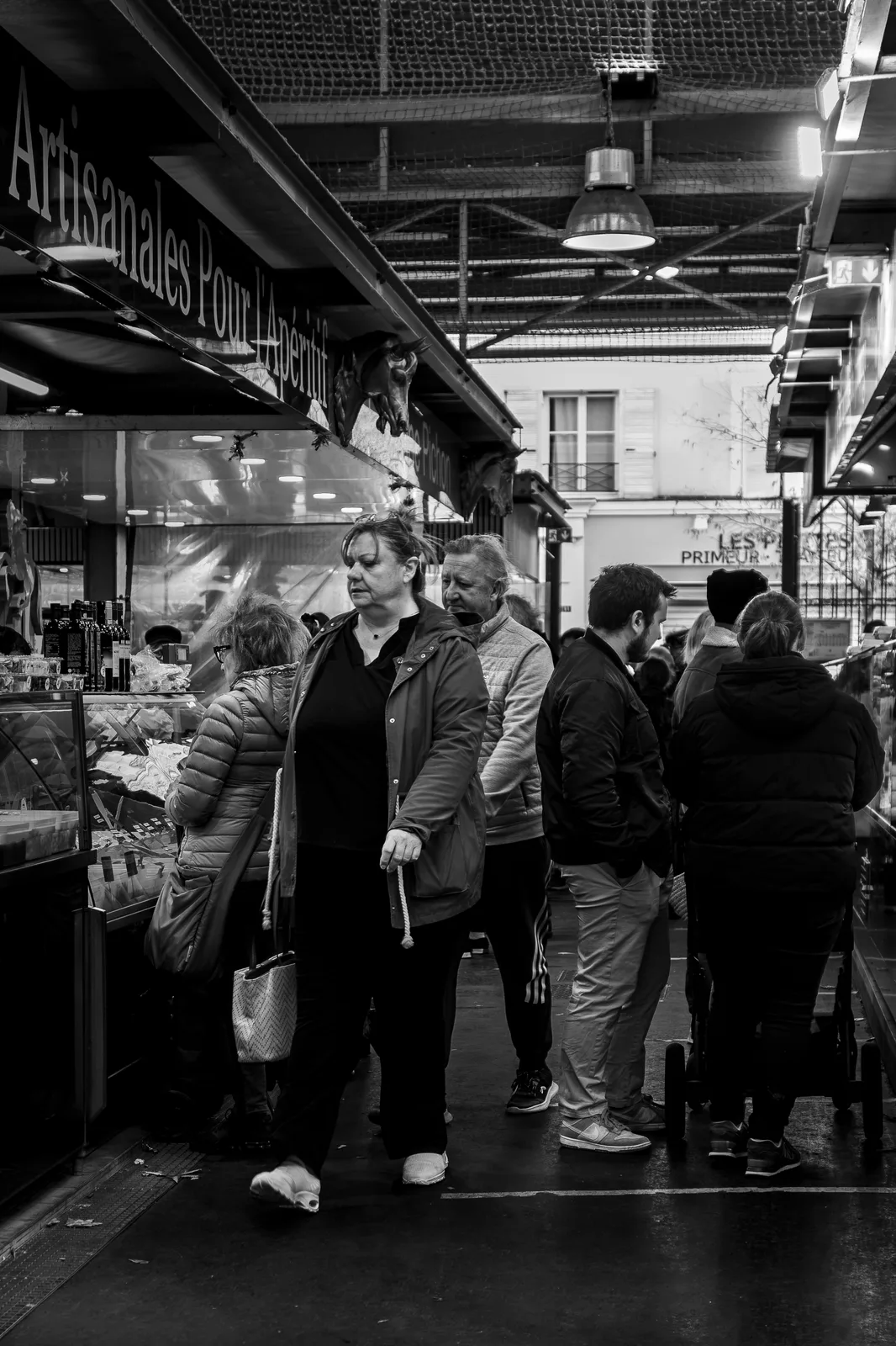 Photographie en noir et blanc d'une allée de marché couvert : une femme marche devant un étal vitré, entourée de clients, sous une structure métallique et une lampe suspendue.
