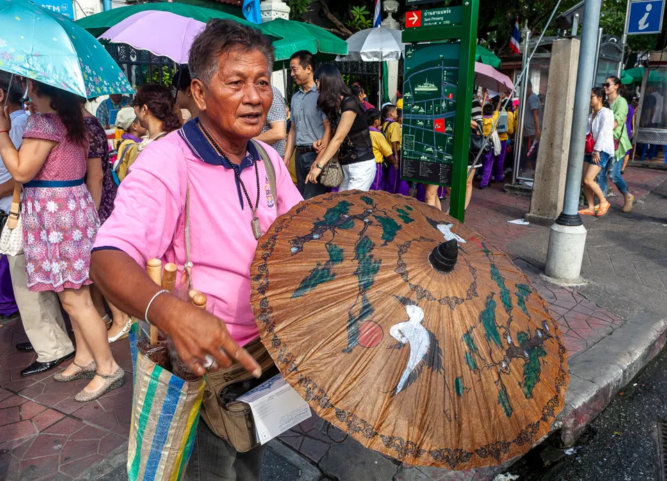 Un homme en polo rose présente une grande ombrelle brune peinte d'oiseaux blancs et de branches, devant un panneau d'information et des passants sous des parapluies.